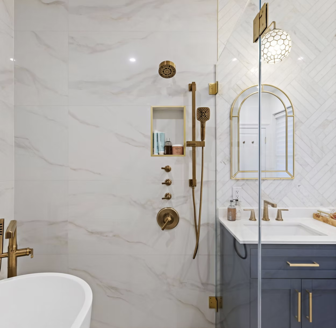 Marble shower with brass fixtures and arched mirror over navy vanity